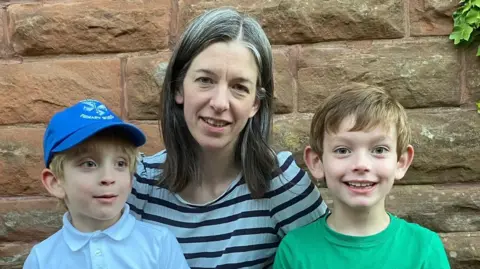 Leigh Leigh is between her two young sons in front of a brick wall 