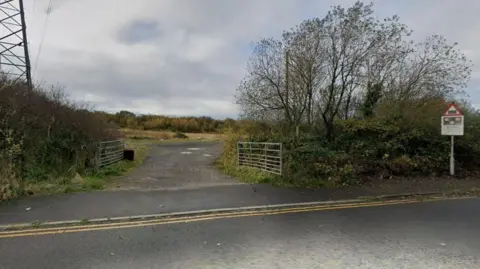 Google View taken from a rural road of an unmade road leading though two open six-bar gates into an area of open scrubland. Trees and shrubs can be seen on either side of the gates, and also beyond the open area.