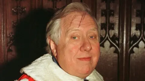 A man with short white hair wearing red and white robes standing in front of dark panelled walls