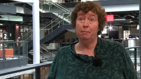 A woman with curly brown hair wearing a green patterned top and standing in front of a staircase.