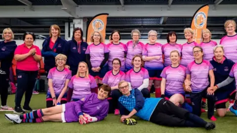 A team of women posing for a picture dressed in pink football kits. They are on a grassy areas in front of seats in a stadium.