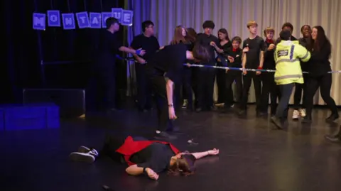 Sophia Seth / BBC A child lying on the floor of a stage. A group of children are standing in the background behind a fake police tape. A blue sign, reading "MONTAGUE" sits in the background.