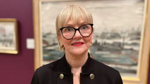 The Chief Executive of The Lowry Julia Fawcett, who has short crop blonde hair and black glasses, smiles for a photograph in front of an LS Lowry painting in an art gallery. 