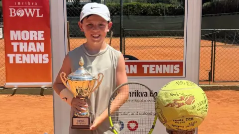 Laura Berridge A young tennis player stands on a clay court holding a trophy, with a racket, bag, and signed tennis ball displayed nearby. Behind the player, banners read “BOWL” and “MORE THAN TENNIS,” placed in front of a fenced court. The scene is brightly lit under the sun, with organised items in the foreground suggesting a recent tournament win.