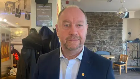 Cefin Campbell, a mad with a short beard and is balding. He is wearing a dark blue suit jacket with a light blue shirt that has the top button undone.