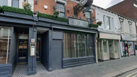 Google The outside of the Dial in Burton, showing a blue framed restaurant front with tall windows and a clock hanging over the front of the restaurant with the words "Dial" painted underneath.