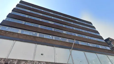 A towering concrete building with five rows of glass windows. The sky is blue.