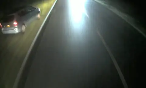 Blurry dashcam screencap of a silver car that's swerved onto a grass verge at the side of a road.