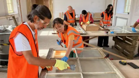 BBC/Tom Ingall Participants wearing high-visibility orange vests, safety glasses and gloves work together to restore historic wooden sash windows in a workshop at Wentworth Woodhouse. Several people scrape old putty and repair window frames at large workbenches, with conservation tools and materials visible throughout the room.