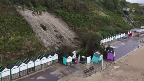 Cluster of beach huts several with turquoise sides and one with purple with tress and debris collapsed from cliffs behind onto them.