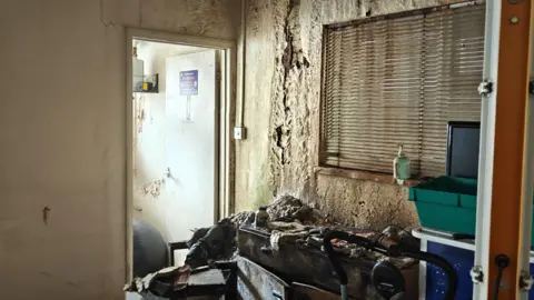 Trevor Snaith/Wycombe Food Hub Severely-damaged room with extensive soot staining and debris, deteriorated walls, damaged furniture and storage units.