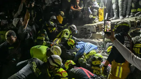 Getty Images A woman is pulled alive from rubble in Columbia by rescue workers wearing neon yellow helmets and vests