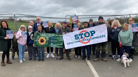 A group of people including men and women stood in a line. They are holding posters, one which reads 'Stop Crofton Development'. In the background is a green field and metal barriers.