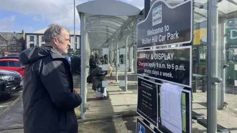 Sir Julian Smith MP Sir Julian Smith is wearing a dark-coloured coat and is reading a sign at Skipton bus station that says 'Welcome to Waller Hill Car Park' with a pay and display sign below. Behind him is a long bus shelter with people waiting for buses and some parked cars.