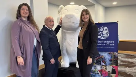 Staffordshire County Council A large white model of a dog, with a heart on a banner around its neck. Three people are stood around the model, one has a hand on its face.
