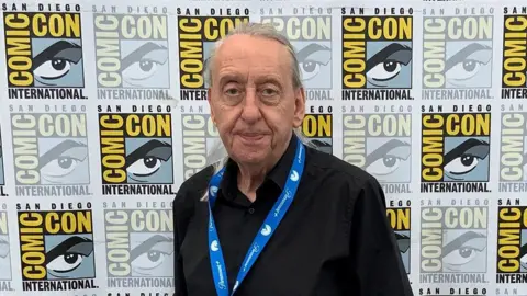 Bryan Talbot A man in a black shirt with long silver hair stands in front of a San Diego Comic Con sign