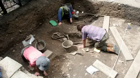 York Archaeology Three men crouched down in the ground during an excavation dig. Skeletal remains are lying in the soil and there are buckets, shovels and planks of wood on the ground.