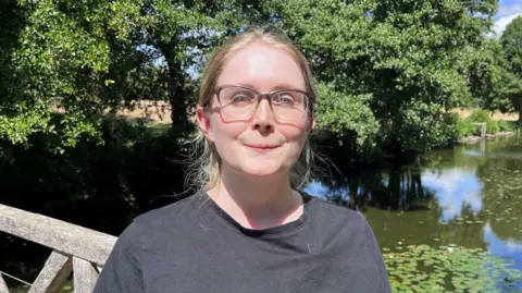 Naomi Collick. She has blonde hair and is wearing a grey T-shirt and spectacles
