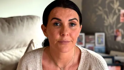A woman looks into the camera with an expressionless face. Her dark hair is tied back. She is wearing a cream cardigan and thin gold necklace.
