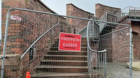 A metal fence placed in front of the steps leading up to Irish Gate Footbridge in Carlisle, with a sign indicating the footpath is closed.