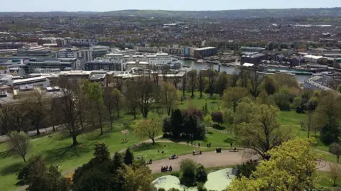 Aerial view of a cityscape with a large park in the foreground, featuring green lawns, trees, walking paths, and people relaxing. A river with docked boats separates the park from a mix of residential and commercial buildings. Rolling hills are visible in the distance under a clear sky