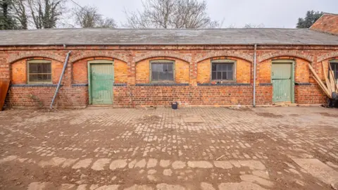 Delapré Abbey Stable buildings, with windows in them, two green doors, an area with stones on the ground and wood to the right. 