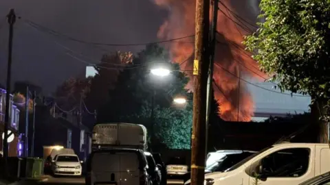 The picture of a housing area has been taken at night-time. There are cars and vans parked on both sides of the street, and telephone poles line the street with trees. In the background is a plume of orange smoke billowing into the sky. 