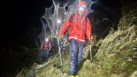 PMRT A group of mountain rescue volunteers walk across rocky, grassy broken ground in the dark. The volunteers are lit up by headtorches. They are wearing red and black waterproofs and use walking poles. 