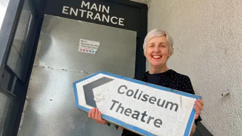 BBC Julie Hesmondhalgh holding up a road sign saying "Coliseum Theatre" in front of the venue's boarded-up main entrance