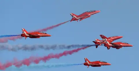 Five Red Arrow Hawk aircraft are flying in a clear blue sky with blue, white and red plumes trailing in the sky behind them.