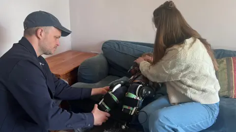 Sam, Asger and Holly in their living room. Together the parents are putting the exoskeleton onto Asger's legs as he lies on a sofa.