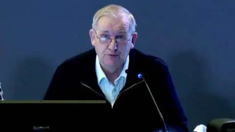 Omagh Bombing Inquiry Jim Sharkey giving evidence to the Omagh Bombing Inquiry.  He has grey hair and glasses and is wearing a light-coloured shirt under a dark-coloured zipped jacket. 