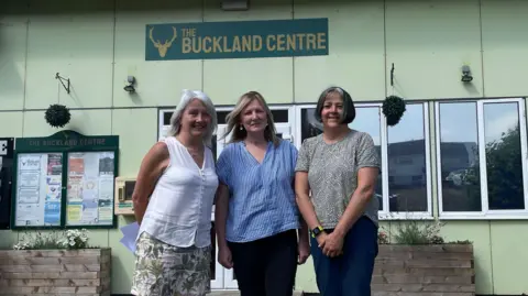 BBC Three middle-aged women stand in front of a pale green building. A sign reading "The Buckland Centre" is on the wall behind them.