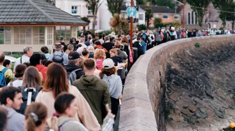 A crowd of people are walking with the seawall to their right and their backs to the camera. It looks like there are more than 100 people walking at dusk.