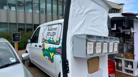 Tom Jackson/BBC A white van with "the VanJarred Refillery" parked outside a building. On the back there are containers of vegan pick-n-mix sweets attached to white awning. 