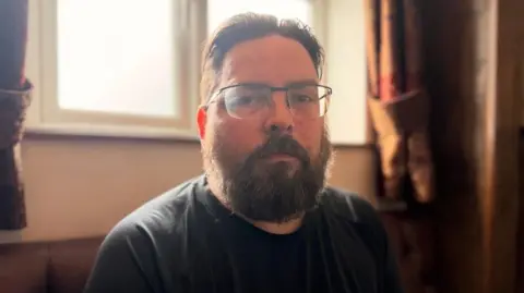 A man with black hair, slicked back and a beard. He is wear black rimmed glasses and looking at the camera with a stern look.