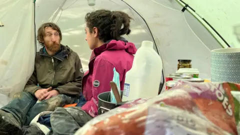 A man and woman sit inside a white tent. The man has long brown hair, a brown beard, a green coat and blue jeans. The woman has black hair tied back, a navy hoodie and a pink coat. There are apples, milk, tins and cups in the foreground.