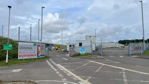 Entrance to Millhill Meadow Recycling Centre with multiple signs, gates, and barriers. The main sign displays the centre's name along with opening times and restrictions. The area is fenced, with streetlights and buildings in the background under a partly cloudy sky.