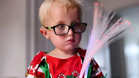 PA Media Five-year-old Harvey Hind with blond hair wearing black glasses in a red, white and green Christmas top with candy canes holds two sensory optic wands which he is looking at. 