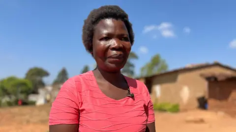 BBC Dorothy Masasa está afuera con una camisa rosa bajo el sol. Al fondo hay un edificio de una sola planta.