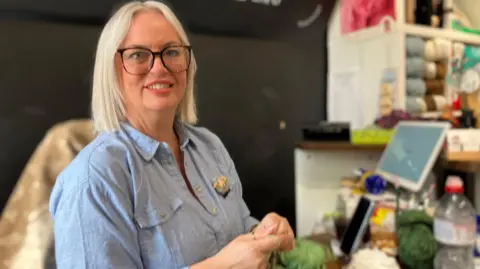 A woman with blonde hair and dark rimmed glasses, wearing a blue denim jacket is smiling. She has wool wrapped around her finger 