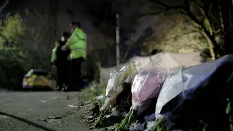 Bouquets of flowers left on Ravenscraig Road, near Ashtons Field in Salford, Greater Manchester, where the remains of a baby were found in a field. Police officers and a police car are in the background of the photograph.