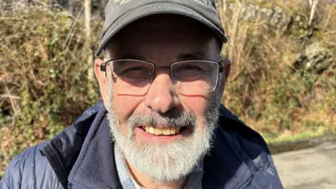 John Urquhart, an older gentleman, with a grey beard and glasses, he wears a navy jacket and a black cap.