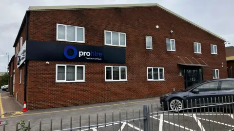 A two-storey red brick building, with a grey car parked in front of it. A large sign wrapped around the corner of the building says "Prohire. The future of fleet"