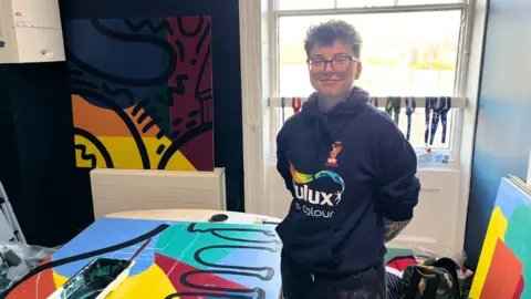 LDRS A woman with short hair and glasses, wearing a Dulux jumper, stands next to a colourful mural.