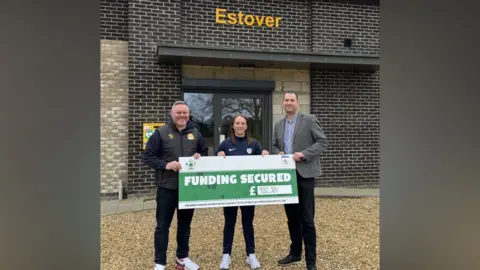 Two men and a woman smile at the camera as they hold a large cardboard cheque that reads: "Funding Secured £982,381". They are standing on gravel and in front of a building. 