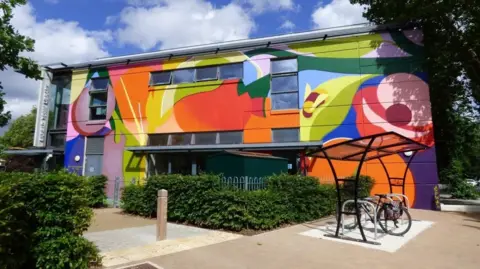 A rectangular library building with one side painted a mixture of bright colours and covered in different shapes. A bike rack with a attached bicycle is located to the right and the library is surrounded by green hedges.