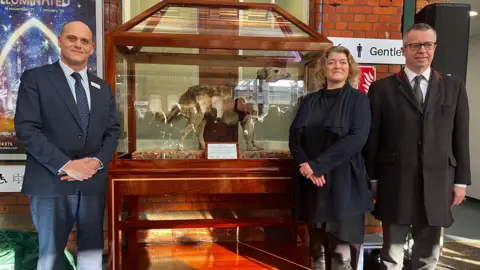 A picture of Station Jim, who is slightly obscured by the reflection of his case, next to the three people who unveiled him on Thursday. Joe Graham from GWR is wearing a dark blue suit, Anna Jones is wearing a blue coat and black skirt, and Tim Knox is wearing a dark coat and grey trousers.