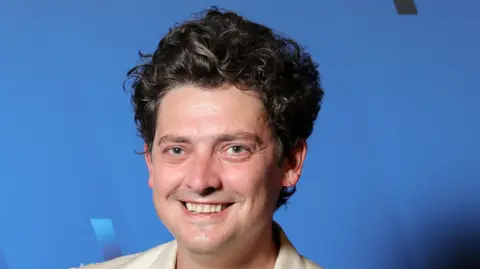 A man with curly brown hair standing in front of a blue background wearing a cream suit jacket anfd smiling at the camera.