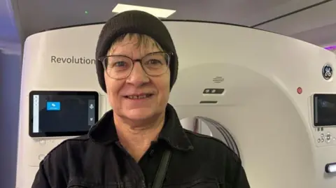 Tracey Markham stood in front of the CT scanner inside the mobile unit. She has a black beanie-style hat on and wears a black jacket.